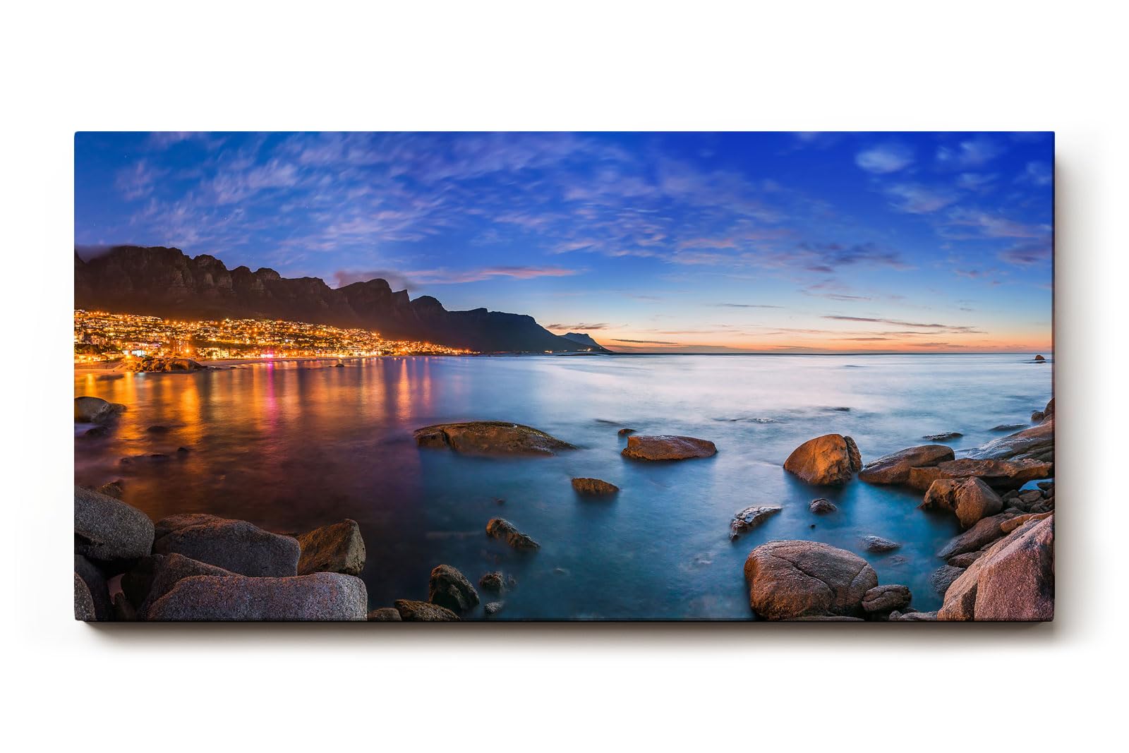 Sinus Art Wandbild 120x80cm Abendliche Szene mit beleuchteter Stadt am Meer, umgeben von Felsen und ruhigem Wasser