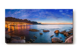 Sinus Art Wandbild 120x80cm Abendliche Szene mit beleuchteter Stadt am Meer, umgeben von Felsen und ruhigem Wasser