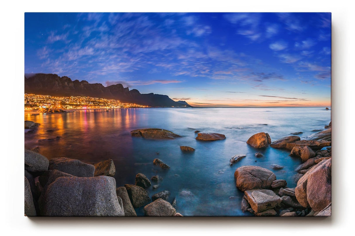 Sinus Art Wandbild 120x80cm Abendliche Szene mit beleuchteter Stadt am Meer, umgeben von Felsen und ruhigem Wasser