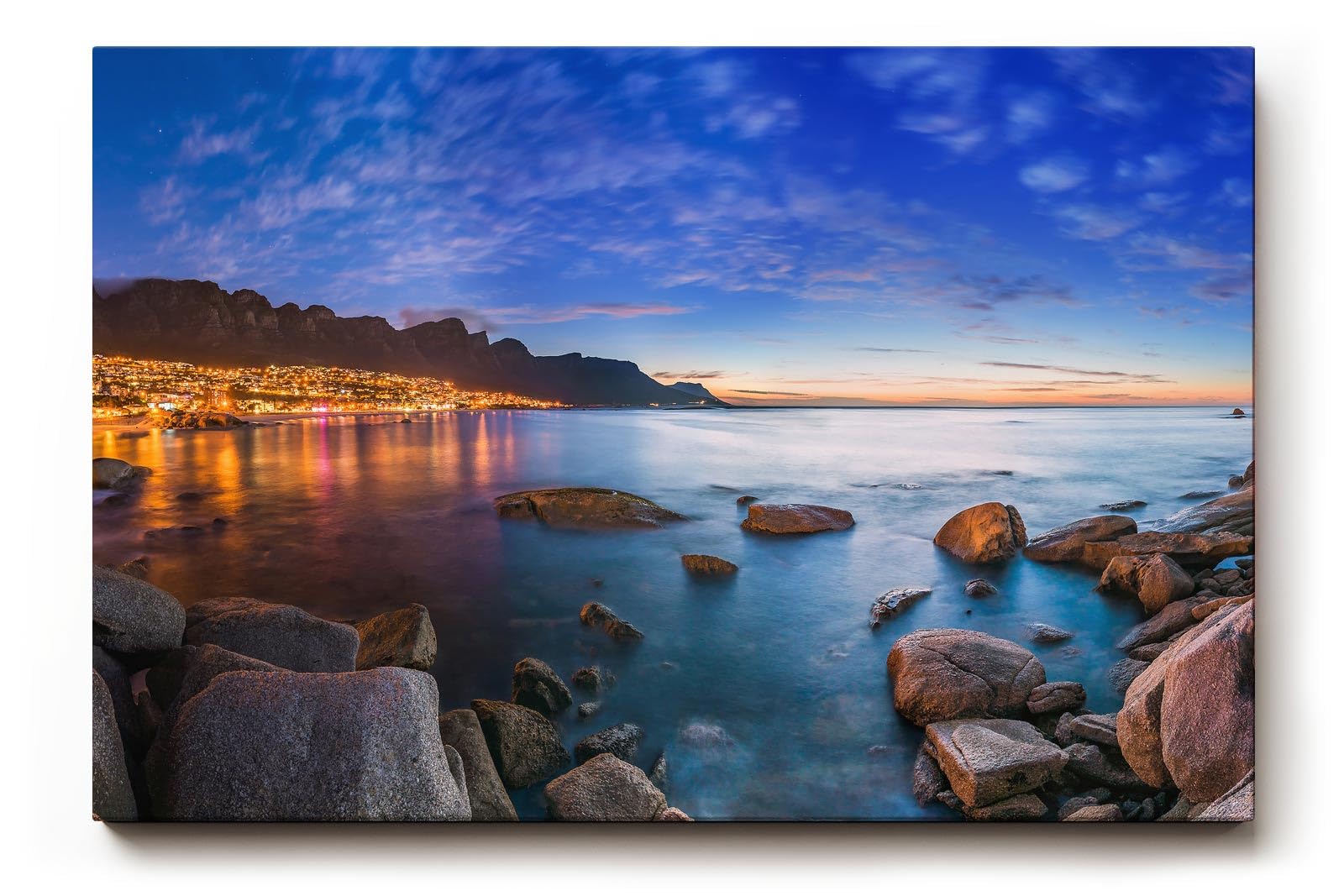 Sinus Art Wandbild 120x80cm Abendliche Szene mit beleuchteter Stadt am Meer, umgeben von Felsen und ruhigem Wasser
