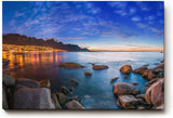 Sinus Art Wandbild 120x80cm Abendliche Szene mit beleuchteter Stadt am Meer, umgeben von Felsen und ruhigem Wasser