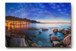Sinus Art Wandbild 120x80cm Abendliche Szene mit beleuchteter Stadt am Meer, umgeben von Felsen und ruhigem Wasser