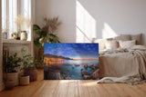 Sinus Art Wandbild 120x80cm Abendliche Szene mit beleuchteter Stadt am Meer, umgeben von Felsen und ruhigem Wasser
